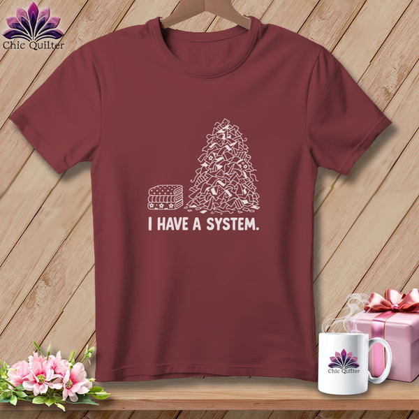Maroon t-shirt with white graphic of fabric scraps and folded fabric, "I HAVE A SYSTEM."