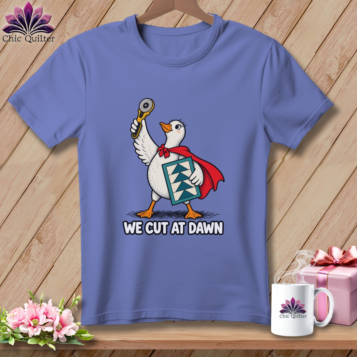 A periwinkle t-shirt features a graphic of a duck wearing a superhero cape, holding a rotary cutter and quilt block, with text reading WE CUT AT DAWN.