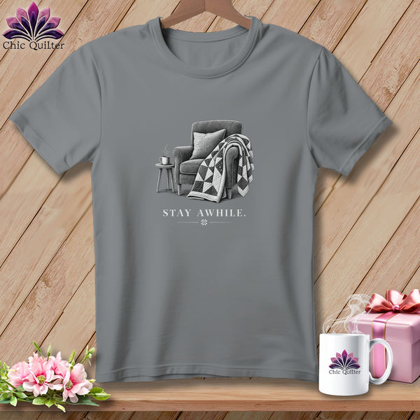 Grey t-shirt with armchair, quilt, and "Stay Awhile" text graphic.