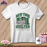 A white t-shirt hanging on a wooden wall displays a green graphic that reads New York Quilter with fabric bolts in a crate.