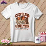 White t-shirt with a Cleveland Quilter graphic featuring a wooden crate filled with fabric rolls.