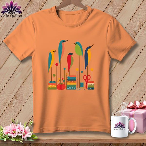 Orange crewneck t-shirt featuring a graphic of colorful stylized birds perched on sewing-themed elements.