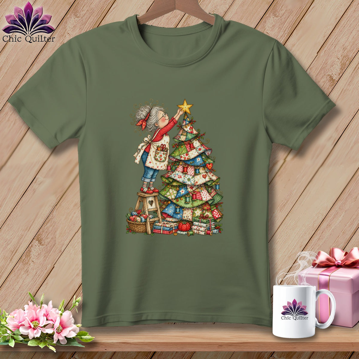 An olive green t-shirt displays a graphic of an elderly woman decorating a patchwork Christmas tree.