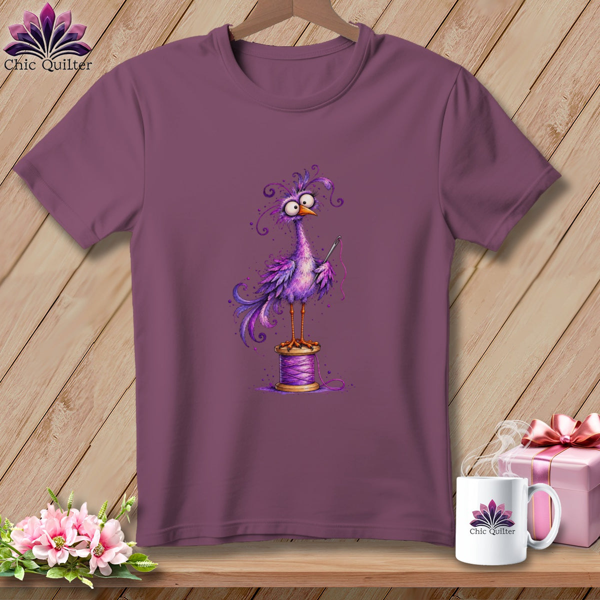 Plum-colored crewneck t-shirt featuring a whimsical purple bird standing on a spool of thread while holding a needle.