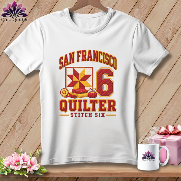 White t-shirt hanging on a wooden wall features a red and yellow San Francisco Quilter graphic with quilting motifs and the number six.