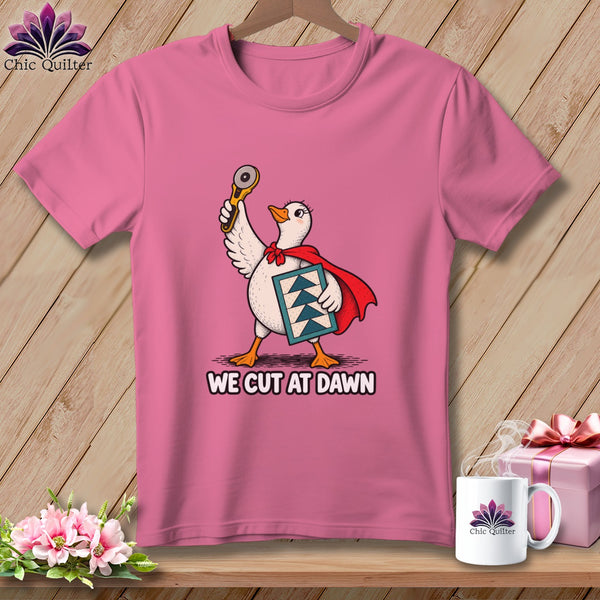 A pink t-shirt features a cartoon goose in a red cape holding a rotary cutter and quilt block, with the text WE CUT AT DAWN.