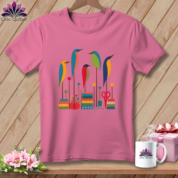 Pink t-shirt displaying a graphic of stylized colorful birds sitting on sewing supplies.