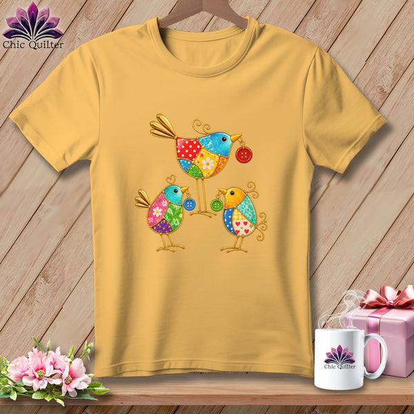 Gold short-sleeve t-shirt featuring a colorful patchwork design of three whimsical metal birds holding buttons.