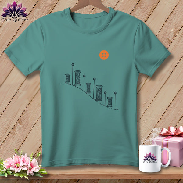 Teal t-shirt with outline of spools of thread and an orange button design.