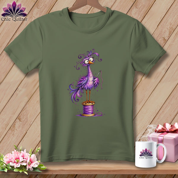 An olive green t-shirt features a whimsical illustration of a purple bird holding a needle while perched atop a spool of thread.