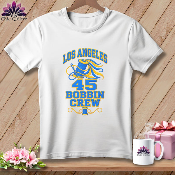 A white short-sleeved t-shirt features blue and yellow text that reads Los Angeles 45 Bobbin Crew with a sewing graphic.