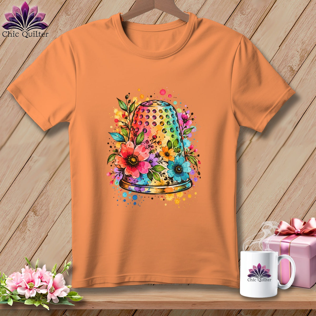 An orange short-sleeved t-shirt features a colorful watercolor thimble graphic surrounded by flowers and paint splatters.