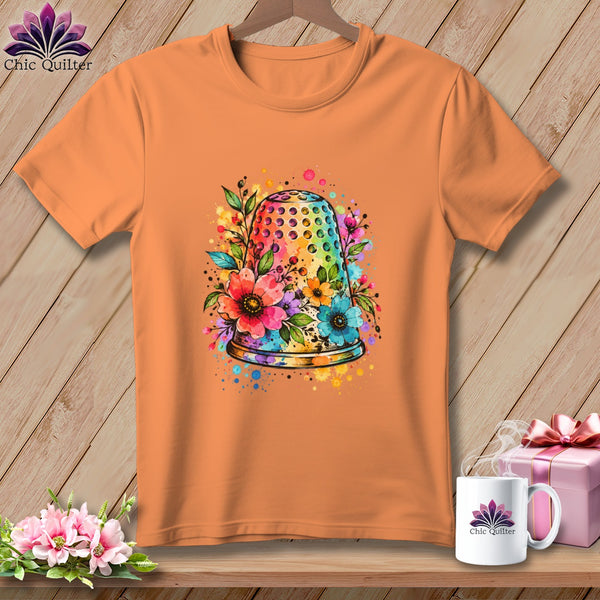 An orange short-sleeved t-shirt features a colorful watercolor thimble graphic surrounded by flowers and paint splatters.