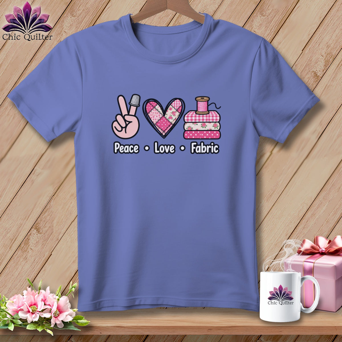 A periwinkle t-shirt features a graphic reading Peace Love Fabric, with a thimble, quilted heart, and folded fabrics.