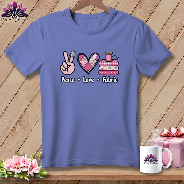 A periwinkle t-shirt features a graphic reading Peace Love Fabric, with a thimble, quilted heart, and folded fabrics.