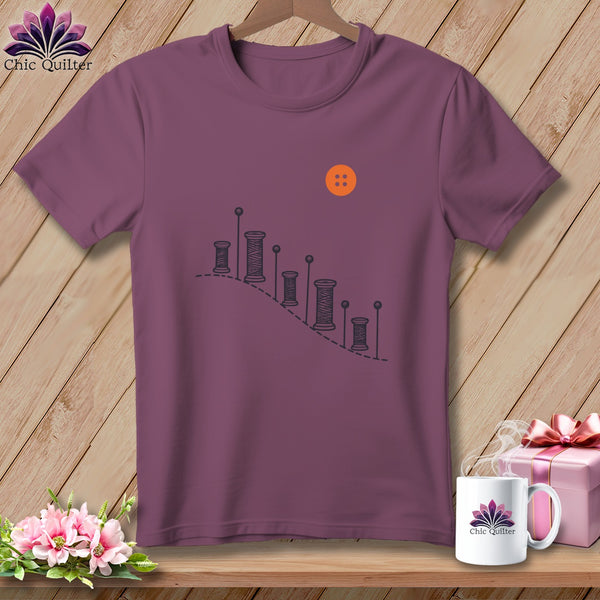 Plum t-shirt with illustration of thread spools, pins, and an orange button.