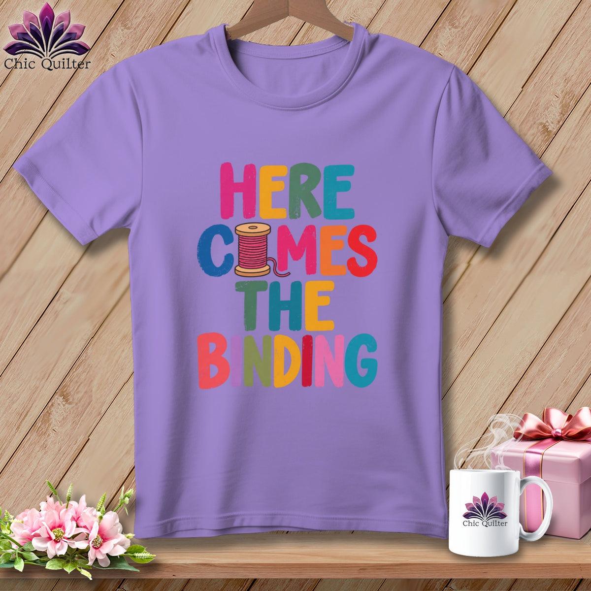 A purple crewneck t-shirt featuring colorful block text reading "Here comes the binding" with a spool of pink thread.