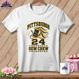 A white t-shirt hanging on a wooden background features a black and yellow graphic with text reading Pittsburgh 24 Sew Crew.