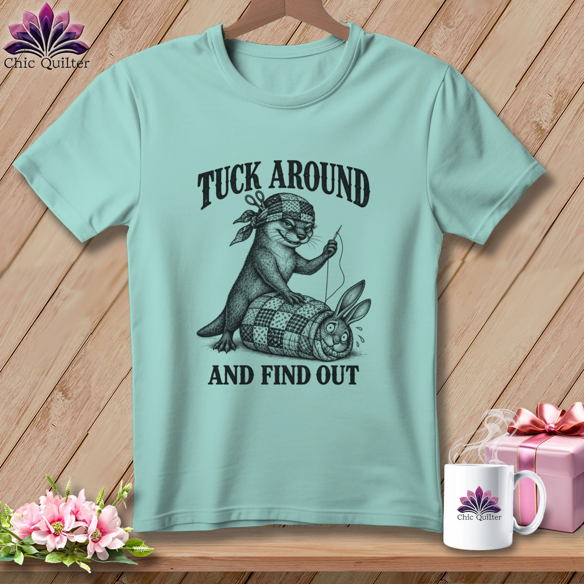 Light blue t-shirt featuring an otter sewing a quilt wrapped around a startled rabbit, with text reading TUCK AROUND AND FIND OUT.