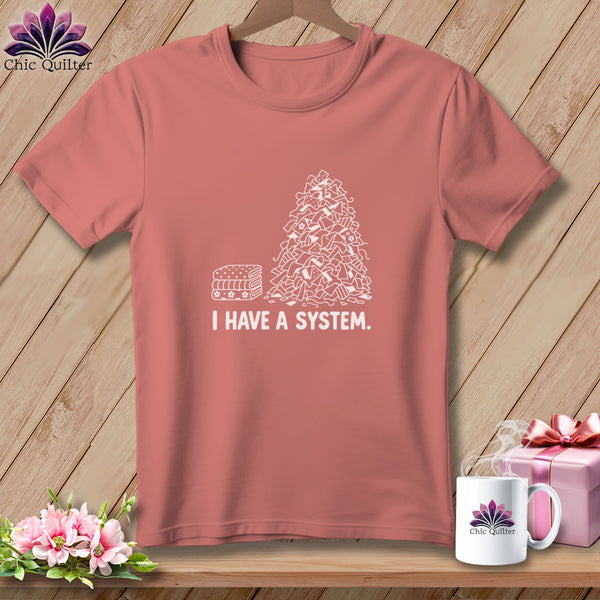 Coral t-shirt with white graphic of stacked fabric and pile of fabric scraps, reads "I HAVE A SYSTEM."