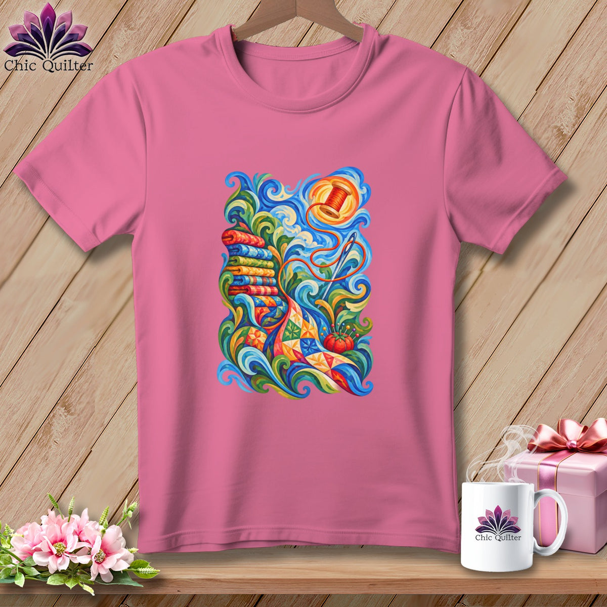 Pink t-shirt featuring a colorful, swirling illustration of quilting supplies including fabric bolts, a needle, and thread.