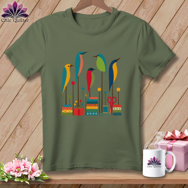 Olive green t-shirt featuring a graphic print of colorful stylized birds perched on sewing-themed elements.