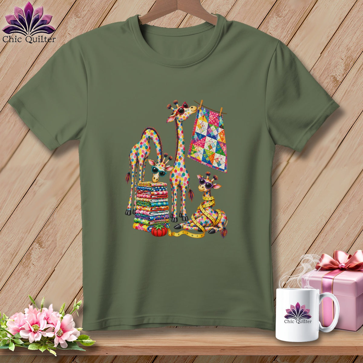 An olive green t-shirt features a colorful graphic of patchwork giraffes, folded fabrics, and quilting tools.