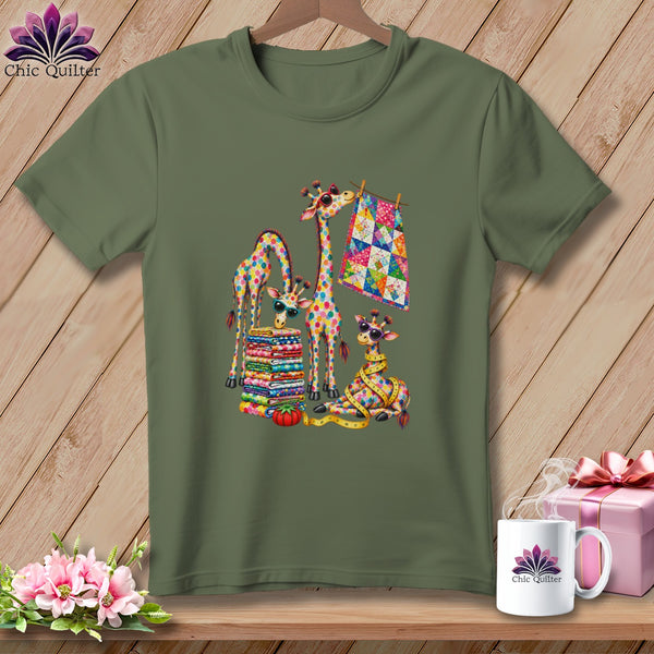 An olive green t-shirt features a colorful graphic of patchwork giraffes, folded fabrics, and quilting tools.