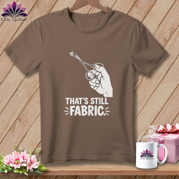 Brown t-shirt with white graphic of hand holding scissors cutting fabric and text "THAT'S STILL FABRIC".