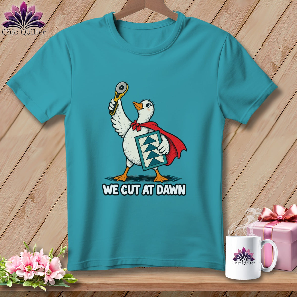 Turquoise t-shirt featuring a graphic of a duck superhero holding a rotary cutter and quilt block, with text reading WE CUT AT DAWN.