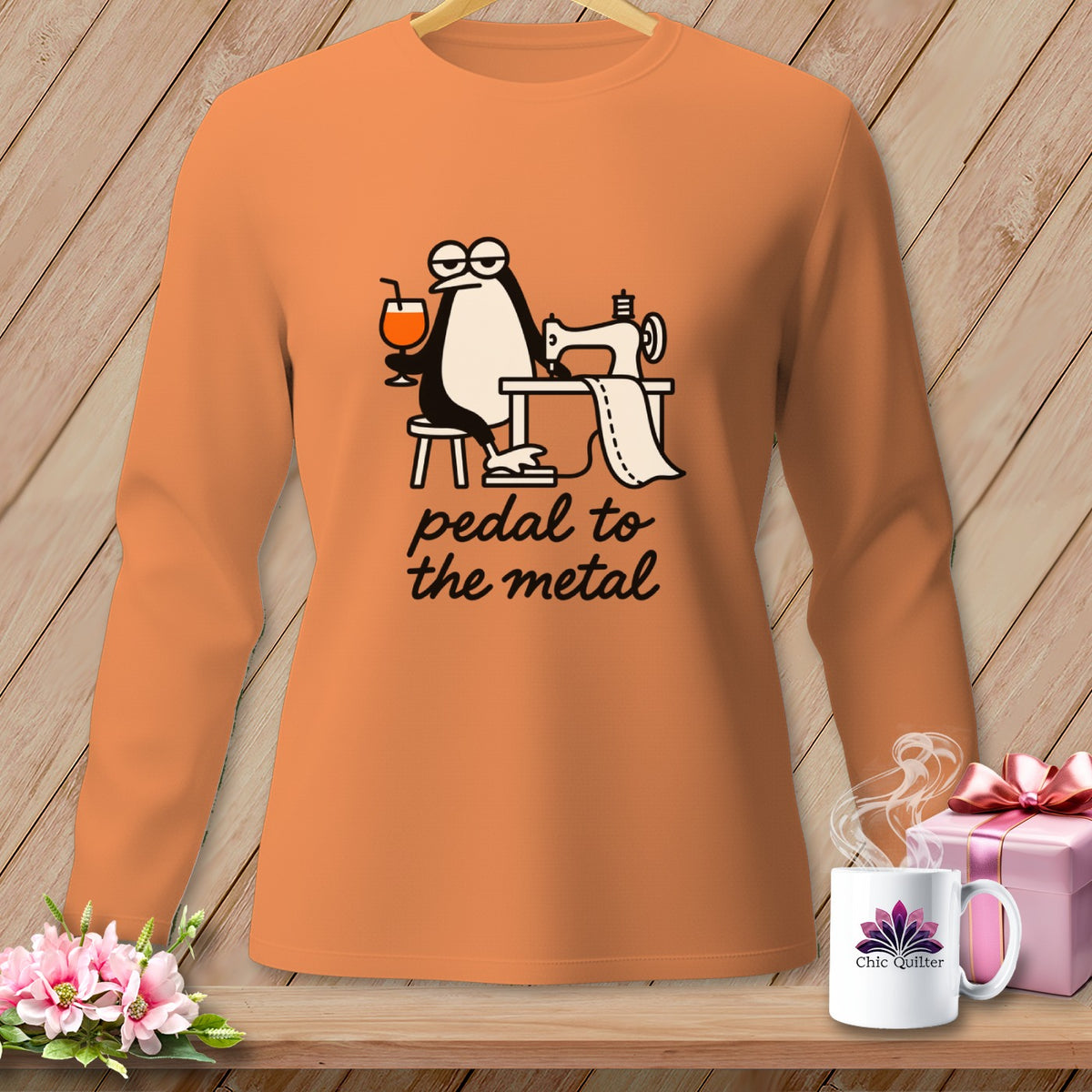 Orange long-sleeve shirt with a cartoon penguin at a sewing machine and the text "pedal to the metal".