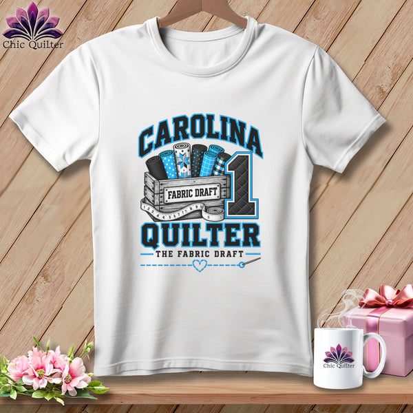 A white short-sleeve t-shirt with a blue and black graphic featuring fabric bolts, a crate, and text reading CAROLINA QUILTER.
