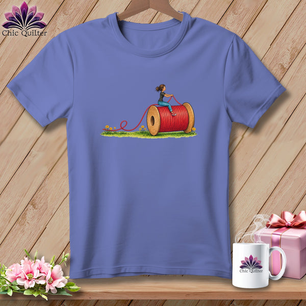 Periwinkle t-shirt on a wooden hanger with a graphic of a woman riding a giant red spool of thread.