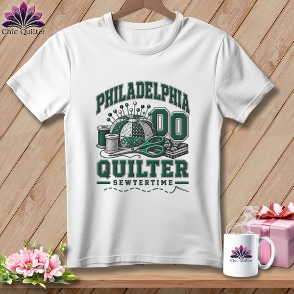 A white t-shirt hanging on a wooden background, featuring green text reading Philadelphia Quilter.