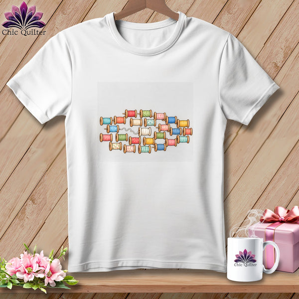 A white short-sleeved t-shirt features a colorful graphic of stacked thread spools on a wooden background.