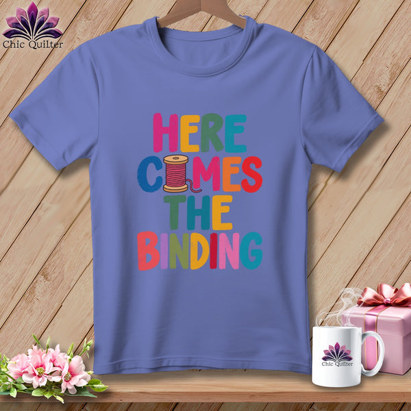 A purple t-shirt with multicolored text reading "HERE COMES THE BINDING" and a pink spool graphic.
