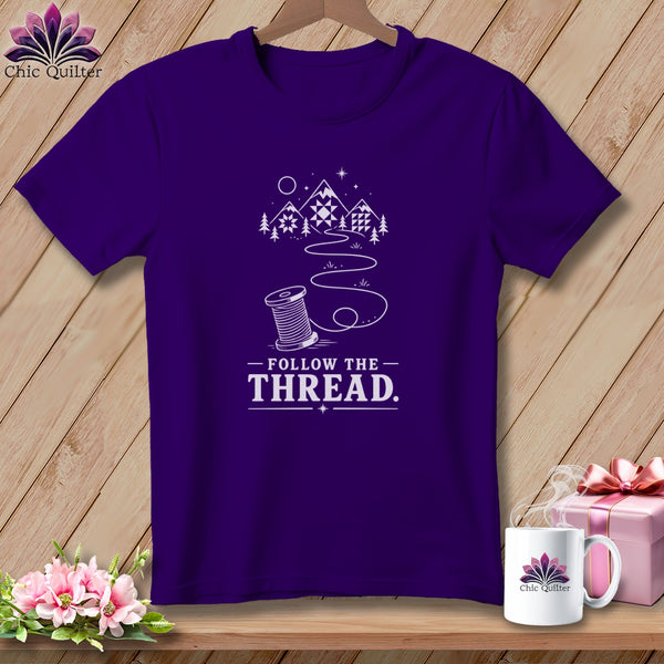 Purple t-shirt with mountains, trees, and a spool of thread design, reading "Follow the Thread."