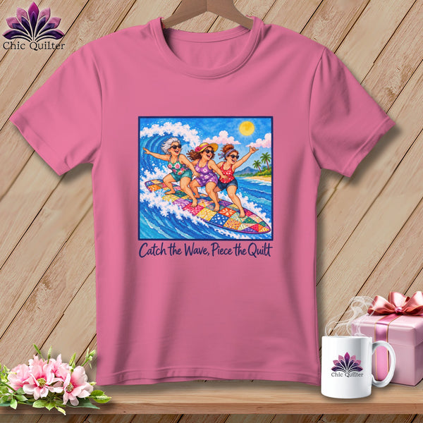 Pink t-shirt featuring an illustration of three women surfing together on a colorful quilt-patterned surfboard.