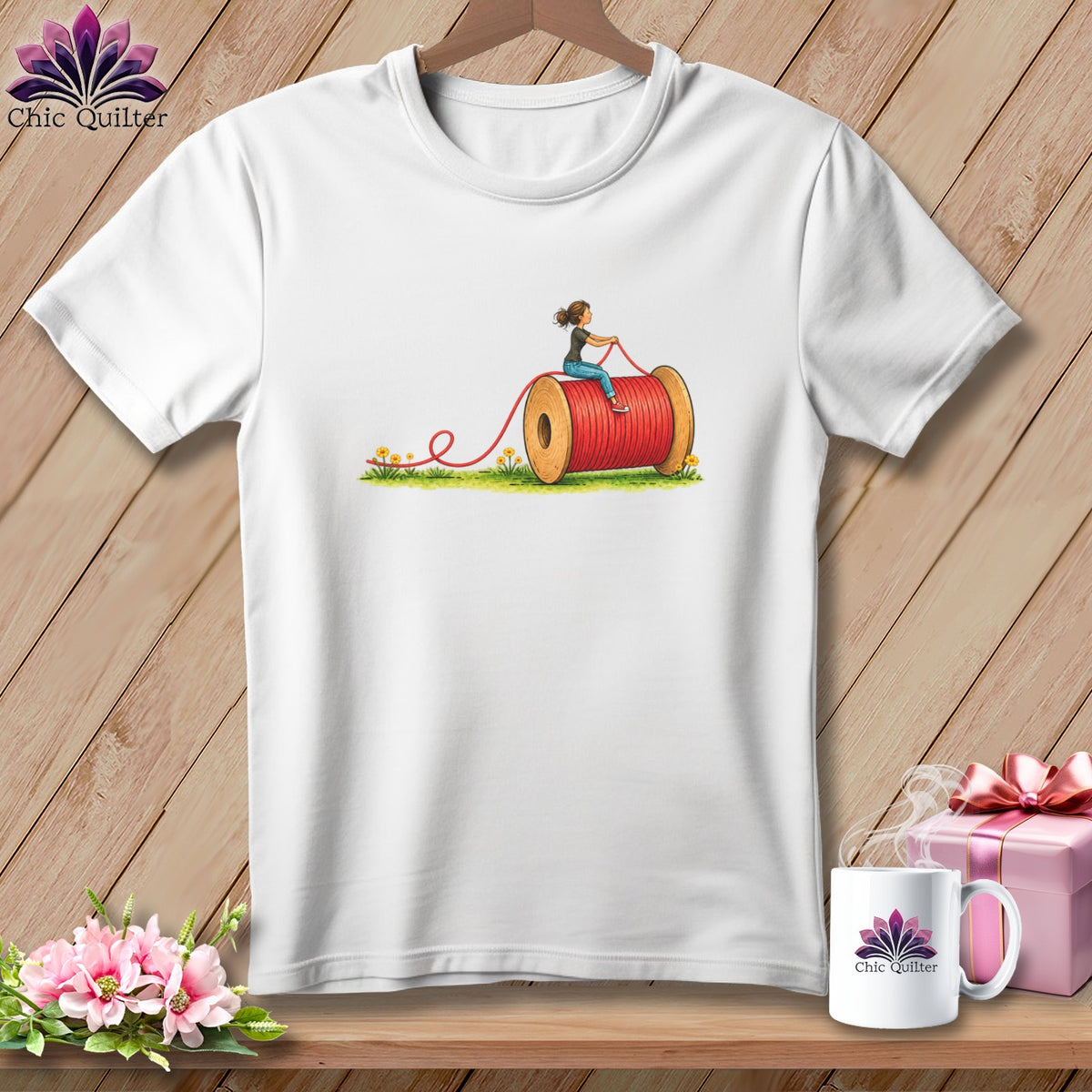A white short-sleeved t-shirt features a graphic of a woman riding on a large red spool of thread.