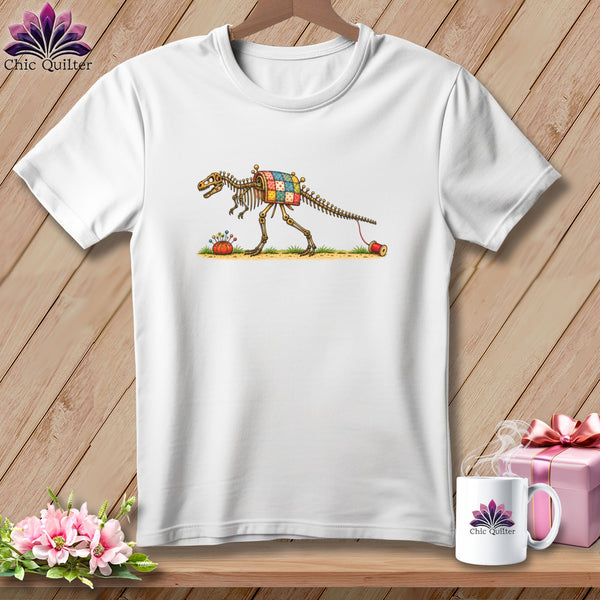 White t-shirt featuring a cartoon dinosaur skeleton wearing a patchwork quilt and pulling a thread spool.