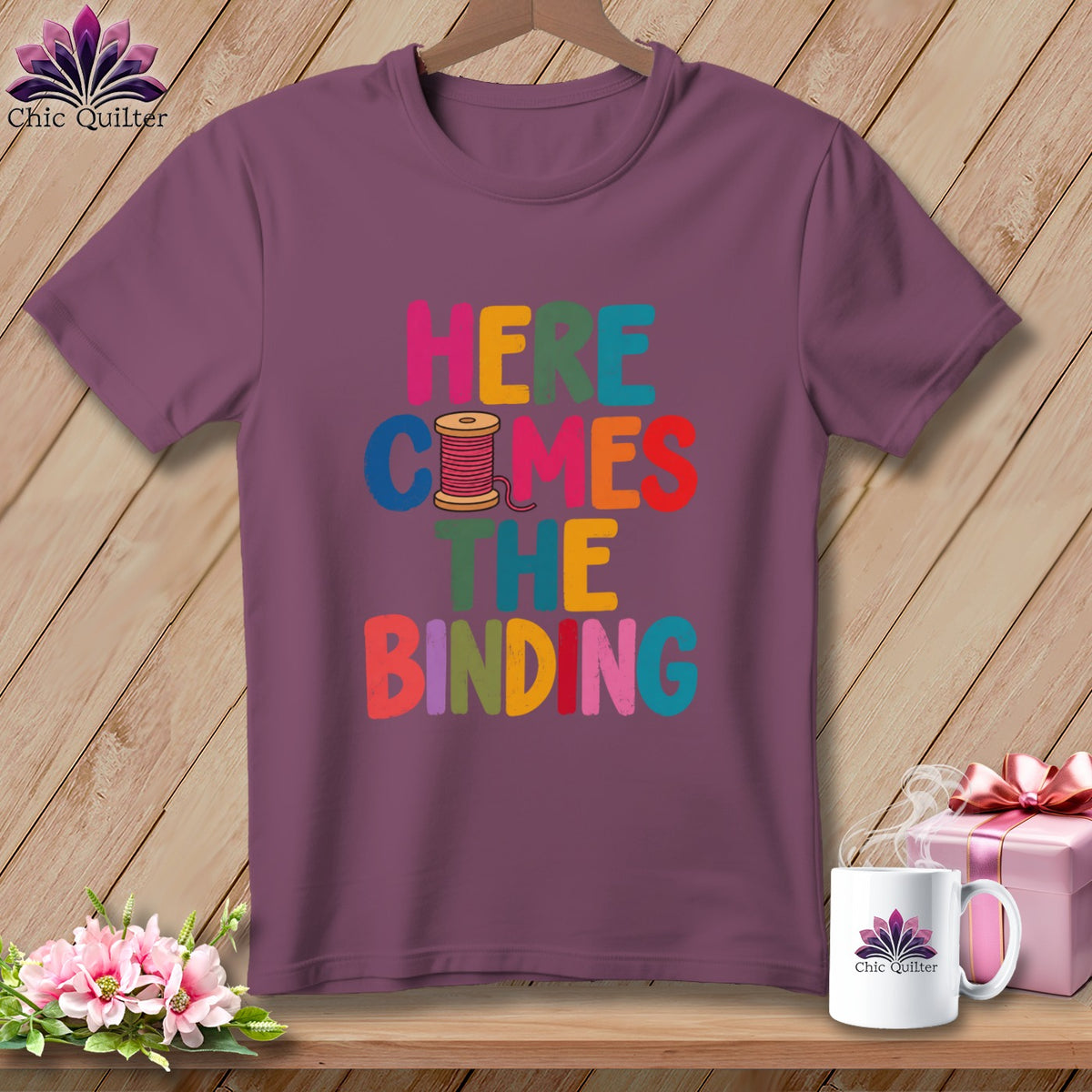A mauve t-shirt on a wooden hanger with the colorful text "HERE COMES THE BINDING".