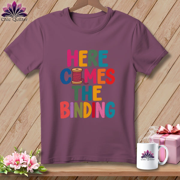 A mauve t-shirt on a wooden hanger with the colorful text "HERE COMES THE BINDING".