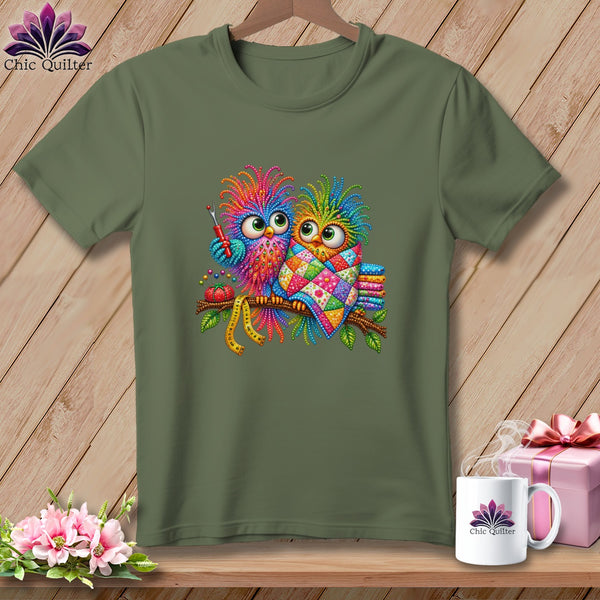 Olive green crewneck t-shirt featuring two colorful, patterned owls perched on a branch, holding sewing tools.