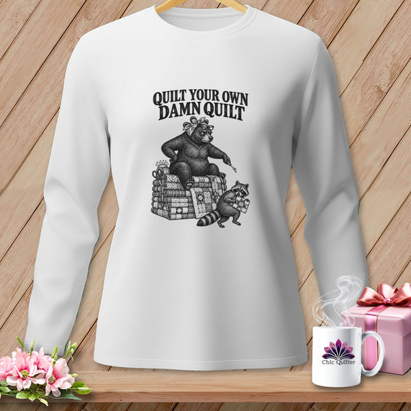 A white long-sleeved shirt featuring a graphic of a bear sitting on quilts with the text QUILT YOUR OWN DAMN QUILT.