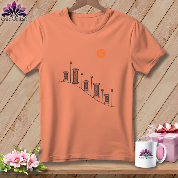Peach t-shirt with a graphic of spools of thread and a button.