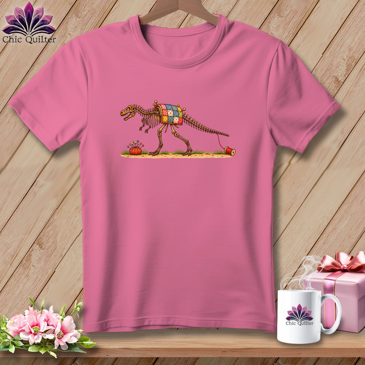 A pink crewneck t-shirt featuring a dinosaur skeleton wearing a colorful patchwork quilt on its back.