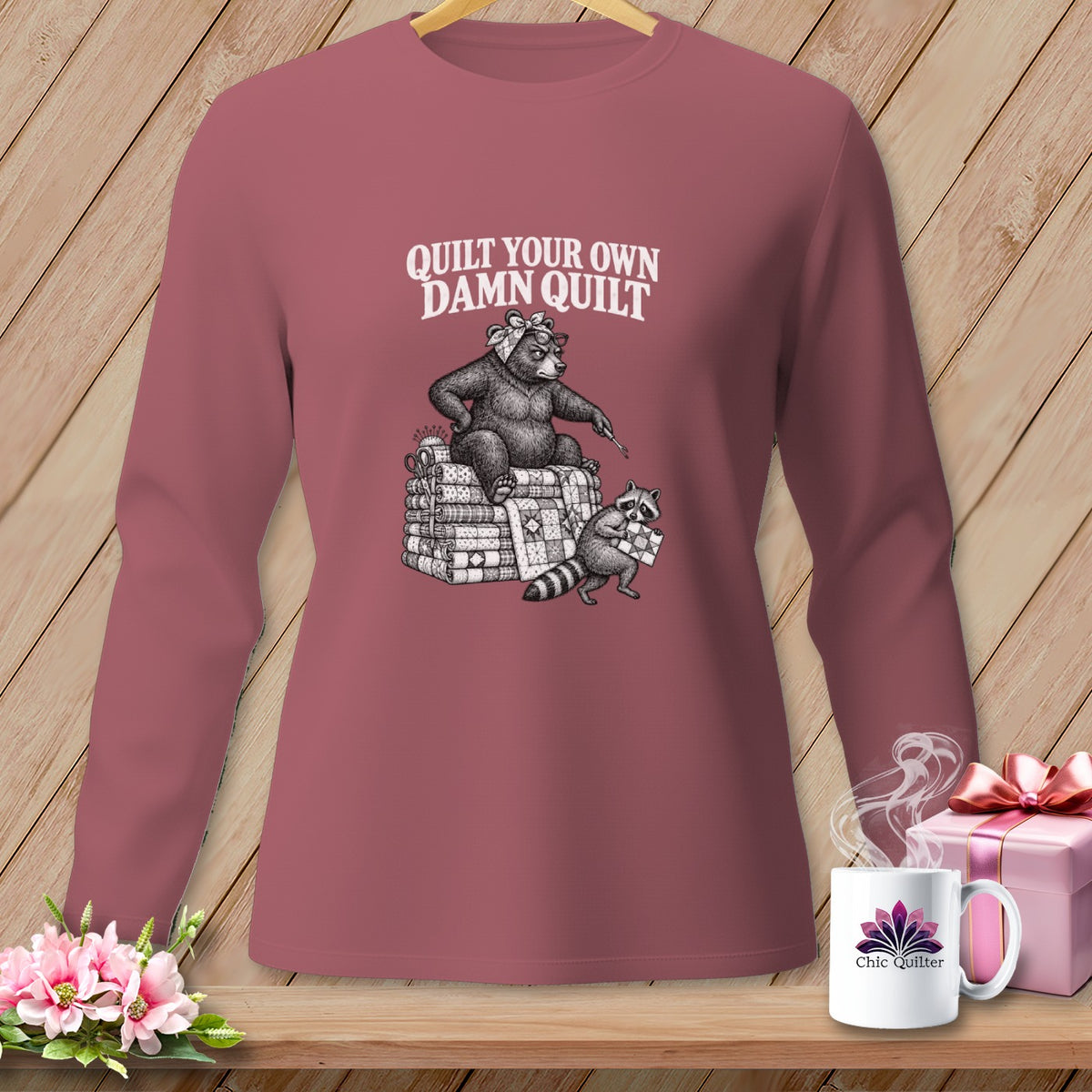 Mauve long-sleeve t-shirt with a vintage illustration of a bear and raccoon on stacked quilts, featuring text reading Quilt Your Own Damn Quilt.