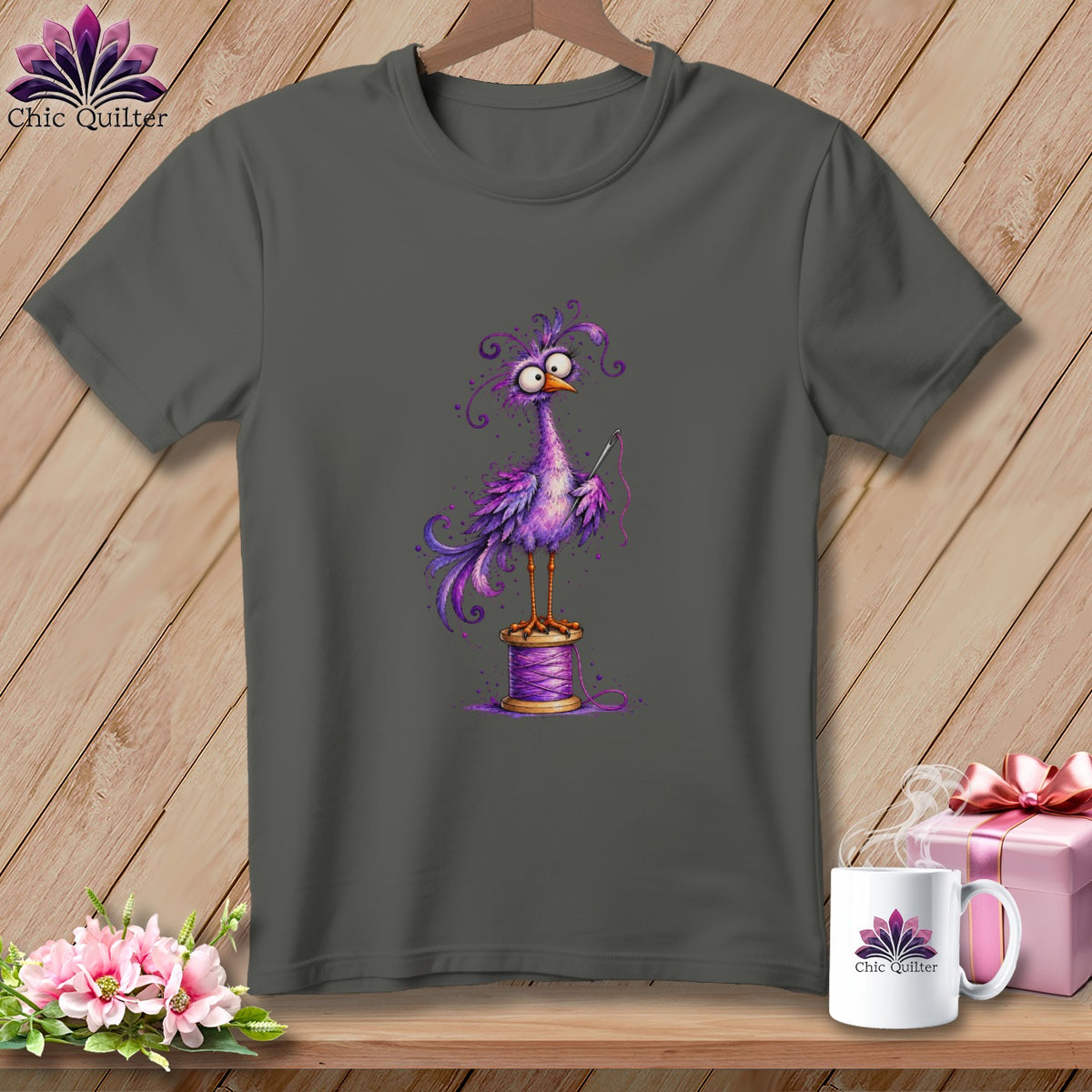 Charcoal gray t-shirt featuring a whimsical purple bird holding a needle while standing on a spool of thread.