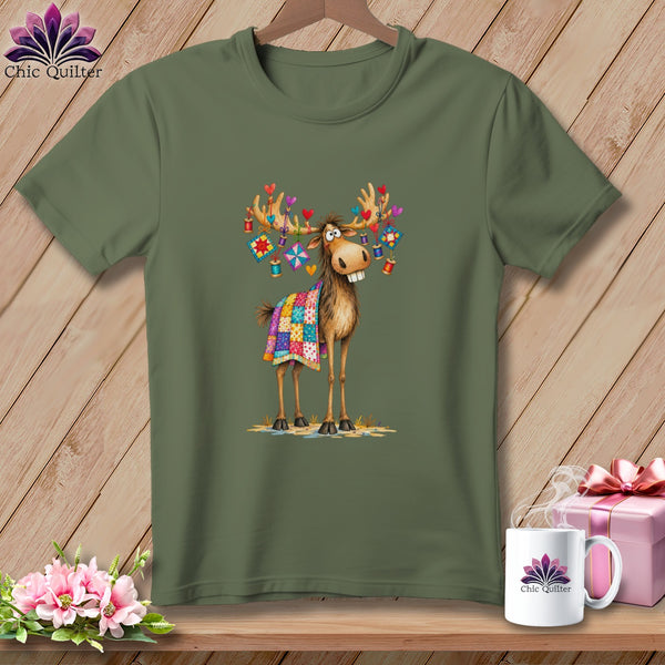 An olive green t-shirt displays an illustration of a cartoon moose wearing a colorful patchwork quilt and hanging decorations on its antlers.