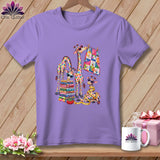 A lavender t-shirt featuring a colorful patchwork giraffe illustration, displayed on a wooden background with a mug and gift.